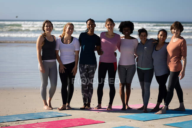Frontansicht einer multiethnischen Gruppe von Freundinnen, die an einem sonnigen Tag die gemeinsame Zeit am Strand genießen, hinter den Yogamatten stehen, Sportkleidung tragen, lächeln, sich umarmen. — Stockfoto
