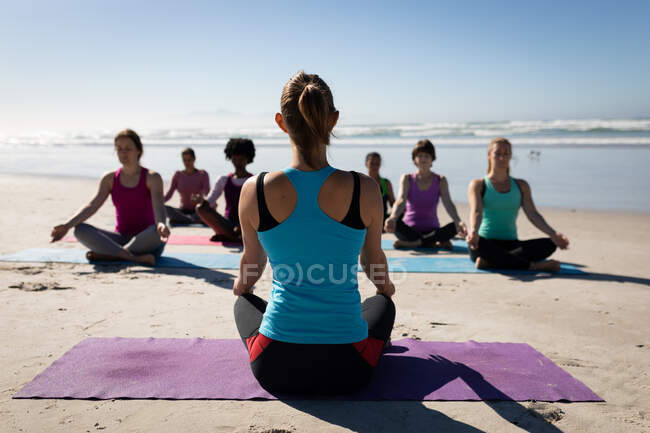 Vista trasera de la mujer caucásica, vestida con ropa deportiva, sentada en una esterilla de yoga, practicando yoga con un grupo de amigas multiétnicas sentadas frente a ella en la playa soleada. - foto de stock