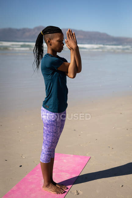 Вид сбоку афроамериканки в пятнистой одежде, стоящей на розовом коврике для йоги с руками в позе йоги на солнечном пляже. — стоковое фото