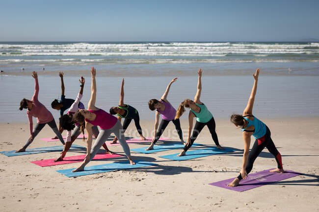 Вид сбоку на многоэтническую группу подруг, наслаждающихся упражнениями на пляже в солнечный день, практикующих йогу, стоящих в позе йоги. — стоковое фото