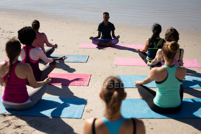 Vista posteriore di un gruppo multietnico di amiche che si esercitano su una spiaggia in una giornata di sole, praticano yoga, si siedono in posizione yoga, meditano. — Foto stock