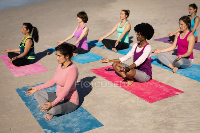 Vista lateral de un grupo multiétnico de amigas disfrutando de hacer ejercicio en una playa en un día soleado, practicar yoga, sentarse en posición de yoga, meditar. - foto de stock