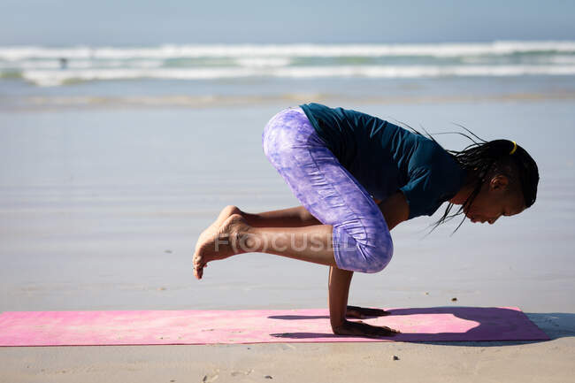 Vista lateral de uma mulher atrativa afro-americana, vestindo roupas esportivas, praticando ioga, de pé em suas mãos em posição de ioga, na praia ensolarada. — Fotografia de Stock