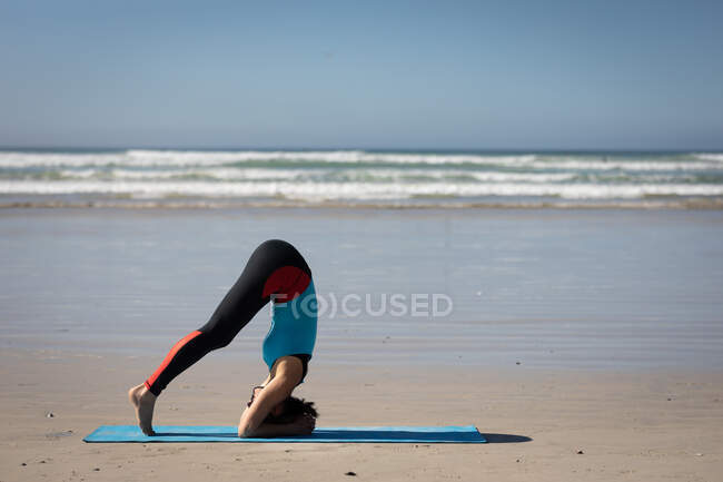 Vista lateral de uma mulher branca atraente, vestindo roupas esportivas, praticando ioga, em pé na posição de ioga, preparando-se para ficar de cabeça, na praia ensolarada. — Fotografia de Stock