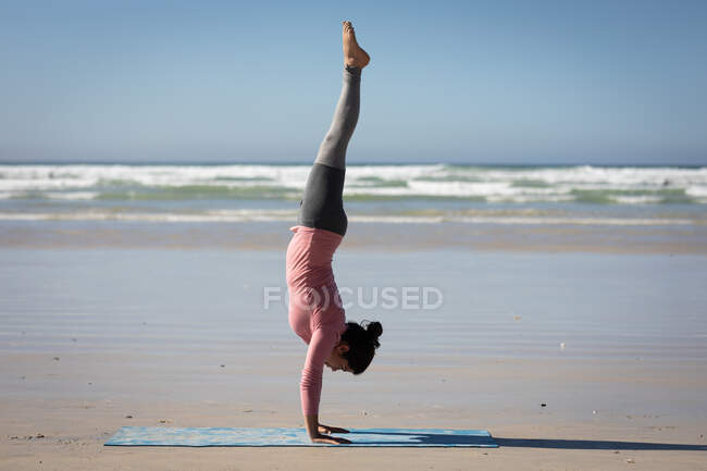 Vista lateral de uma mulher branca atraente, vestindo roupas esportivas, praticando ioga, de pé em suas mãos em posição de ioga, na praia ensolarada. — Fotografia de Stock