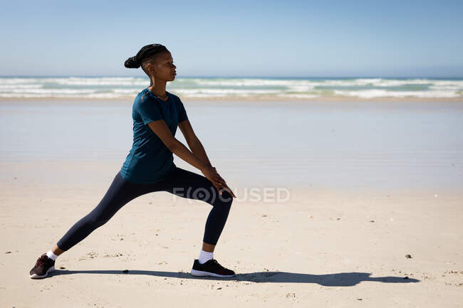 Seitenansicht einer attraktiven afroamerikanischen Frau, die Sportkleidung trägt, Yoga praktiziert, sich in Yogaposition dehnt, am sonnigen Strand. — Stockfoto