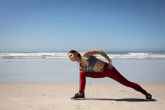 Vista frontale di una donna attraente caucasica, indossando vestiti sportivi, praticando yoga, allungando e appoggiandosi in avanti in posizione yoga, sulla spiaggia soleggiata. — Foto stock