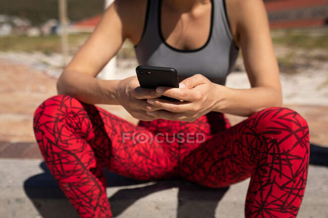 Вид спереду середня секція жінки, одягнена в спортивний одяг, сидить на березі, розслабляється після бігу, тримає смартфон в обох руках, текстовий масаж . — стокове фото