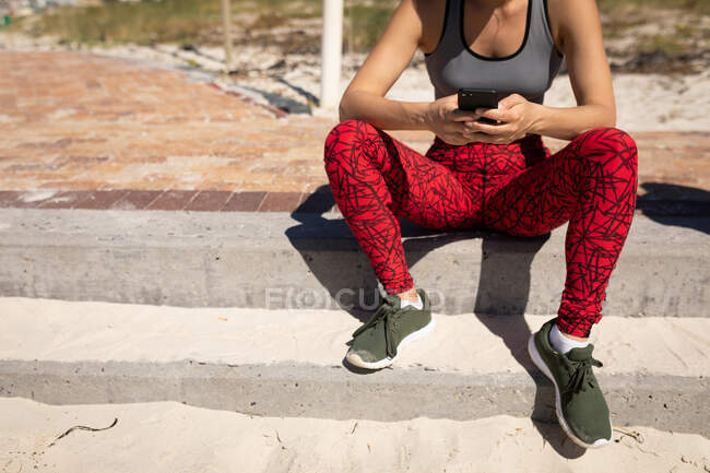 Вид спереду середня секція жінки, одягнена в спортивний одяг, сидить на березі, розслабляється після бігу, тримає свій смартфон в обох руках, текстовий масаж . — стокове фото
