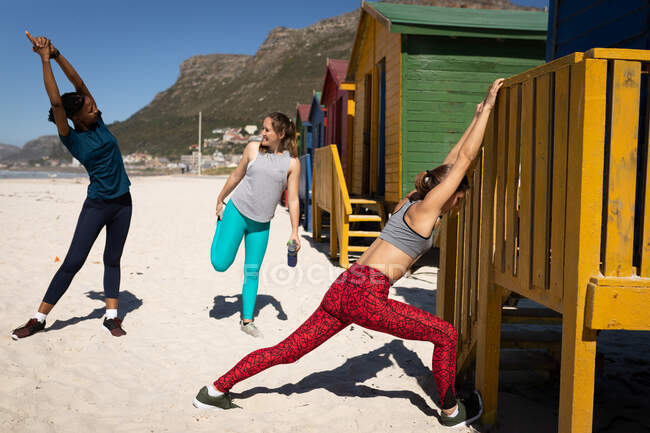 Vue de face d'un groupe multi-ethnique d'amies profitant de l'exercice sur une plage par une journée ensoleillée, s'étirant et se relaxant après le jogging. — Photo de stock