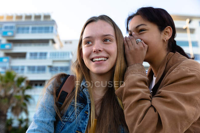 Vista frontal close-up de um caucasiano e uma raça mista meninas desfrutando de tempo pendurado juntos em um dia ensolarado, sorrindo e sussurrando . — Fotografia de Stock