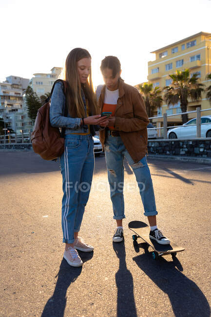 Передній вид закритий Кавказькою і змішаною расою дівчата, які проводять час разом в сонячний день, стоять на вулиці, дівчинка тримає свій смартфон і показує його своєму другу. — стокове фото