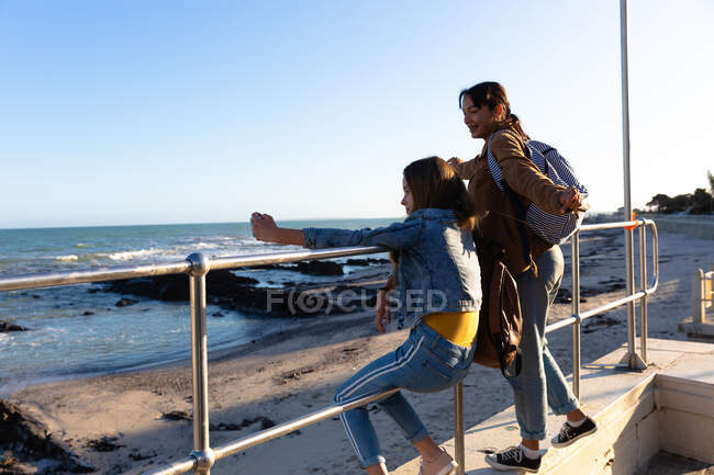 Vue latérale d'une Caucasienne et d'une fille métissée profitant du temps passé ensemble par une journée ensoleillée, fille assise sur une clôture dans une promenade au bord de la mer, portant des sacs à dos, fille prenant selfie d'elle-même et de son amie. — Photo de stock