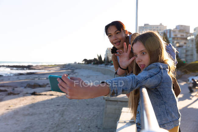 Vista lateral de una caucásica y una mezcla de chicas de raza disfrutando del tiempo juntos en un día soleado, de pie y apoyado en la valla en un paseo marítimo, chica tomando selfie de sí misma y su amiga, haciendo caras divertidas. - foto de stock