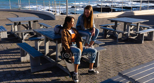 Вид сбоку на кавказку и смешанную расу девушек, наслаждающихся временем, проведенным вместе в солнечный день, сидящих на скамейке на набережной у моря, разговаривающих. — стоковое фото