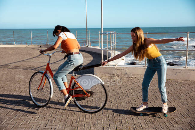 Side view of a Caucasian and a mixed race girls enjoying time hanging out together on a sunny day, playing on promenade by the sea, one girl riding a bike and pulling another on skateboard. — Stock Photo