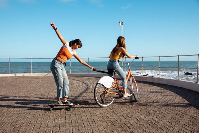 Side view of a Caucasian and a mixed race girls enjoying time hanging out together on a sunny day, playing on promenade by the sea, one girl riding a bike and pulling another on skateboard. — Stock Photo