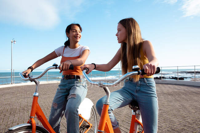 Вид спереди на кавказца и смешанных расовых девушек, веселящихся вместе в солнечный день, играющих на набережной у моря, катающихся на велосипеде, улыбающихся друг другу. — стоковое фото