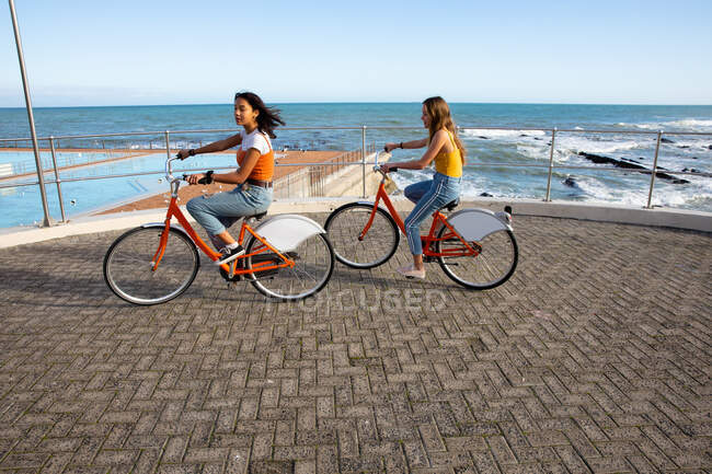 Vista lateral de um caucasiano e uma raça mista meninas desfrutando de tempo pendurado juntos em um dia ensolarado, jogando no passeio marítimo, andando de bicicleta . — Fotografia de Stock