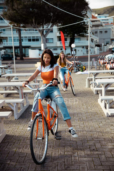 Передній вид на кавказьку і змішану расу дівчат, які проводять час разом у сонячний день, їздять на велосипеді в міському пішохідному районі, посміхаючись. — стокове фото