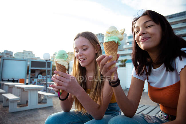 Передній вид на кавказьку і змішану расу дівчат, які проводять час разом у сонячний день, їдять морозиво, сидячи на лавці в міському пішохідному районі, посміхаючись. — стокове фото