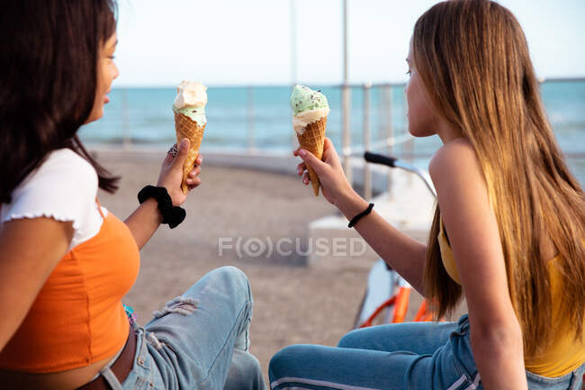 Rückansicht einer kaukasischen und einer gemischten Rasse Mädchen genießen die Zeit zusammen hängen an einem sonnigen Tag, Eis essen, auf einer Bank in einer Promenade am Meer sitzen. — Stockfoto