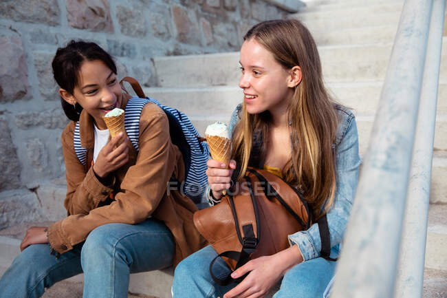 Vorderansicht einer kaukasischen und einer gemischten Rasse Mädchen genießen die Zeit zusammen hängen an einem sonnigen Tag, Eis essen, sitzen auf der Treppe in einer Promenade am Meer. — Stockfoto