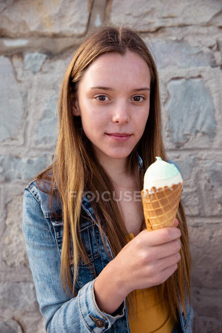 Ritratto di una ragazza caucasica che indossa una giacca di jeans, si gode il tempo passando una giornata di sole, sta vicino al muro, tiene un gelato, guarda dritto verso la macchina fotografica. — Foto stock