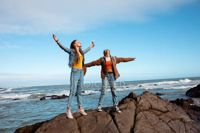 Вид спереди кавказца и смешанных расовых девушек, веселящихся вместе в солнечный день, стоящих с распростертыми на скале руками на пляже. — стоковое фото