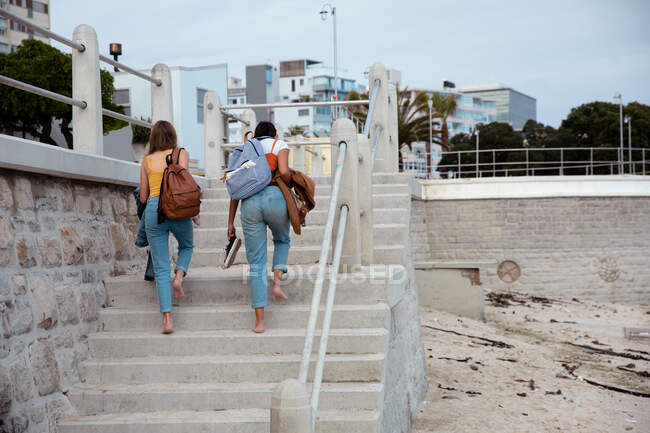 Vista trasera de un caucásico y una raza mixta niñas disfrutando del tiempo juntos en un día soleado, con mochilas, subiendo las escaleras a un paseo marítimo. - foto de stock