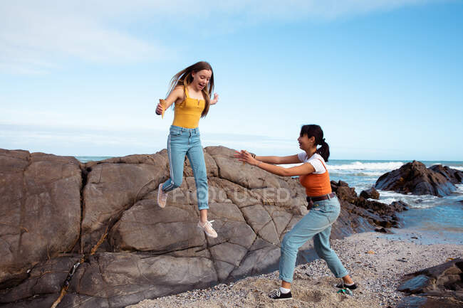 Vista frontal de um caucasiano e uma raça mista meninas desfrutando de tempo pendurado juntos em um dia ensolarado, uma menina pulando de uma rocha na praia, outra tentando pegá-la . — Fotografia de Stock