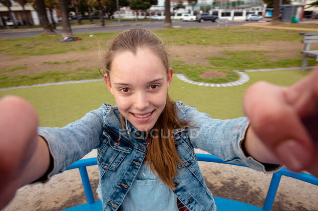 Retrato de una chica caucásica con el pelo largo disfrutando del tiempo pasando el rato en un día soleado en el patio de recreo, tomando selfie de sí misma. - foto de stock