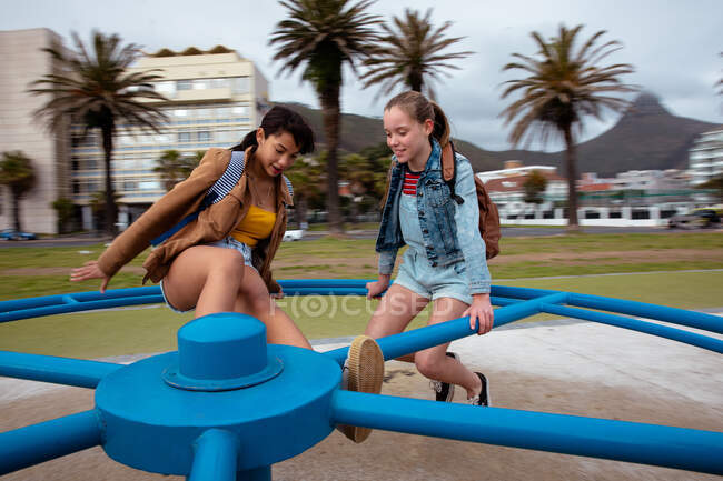 Передній вид на кавказьку і змішану расу дівчат, які проводять час разом в сонячний день на ігровому майданчику, сидячи на веселому круглі, посміхаючись. — стокове фото