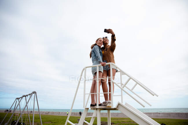 Front view of a Caucasian and a mixed race girls enjoying time hanging out together on a sunny day, standing together on the top on a slide, girl taking selfie of herself and her friend. — Stock Photo