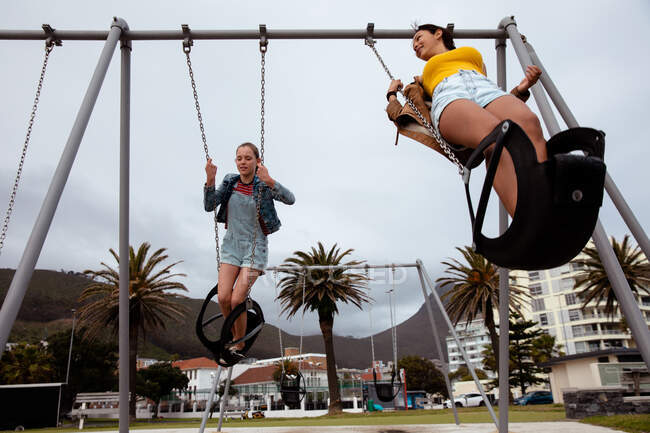 Visão de baixo ângulo de um caucasiano e uma raça mista meninas desfrutando de tempo pendurado juntos em um dia ensolarado, jogando no playground, balançando em um balanço . — Fotografia de Stock