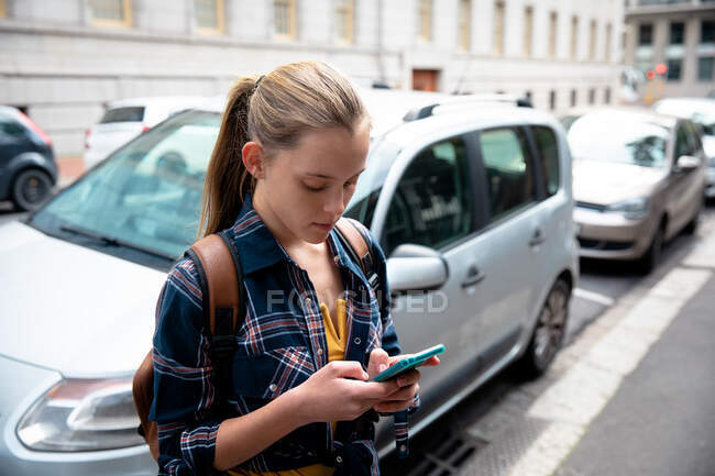 Vue latérale d'une fille caucasienne portant une chemise à carreaux, profitant du temps passé à traîner par une journée ensoleillée, debout sur le trottoir, tenant son smartphone et envoyant un message de massage. — Photo de stock