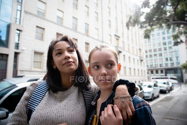 Передній вид на кавказьку і змішану расу дівчат, які проводять час разом в сонячний день, стоячи на тротуарі, обіймаючи. — стокове фото