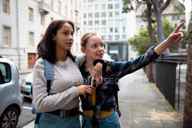 Вид спереди кавказца и смешанных расовых девушек, наслаждающихся временем, проведенным вместе в солнечный день, стоящих на тротуаре, обнимающихся, указывающих на девушку. — стоковое фото