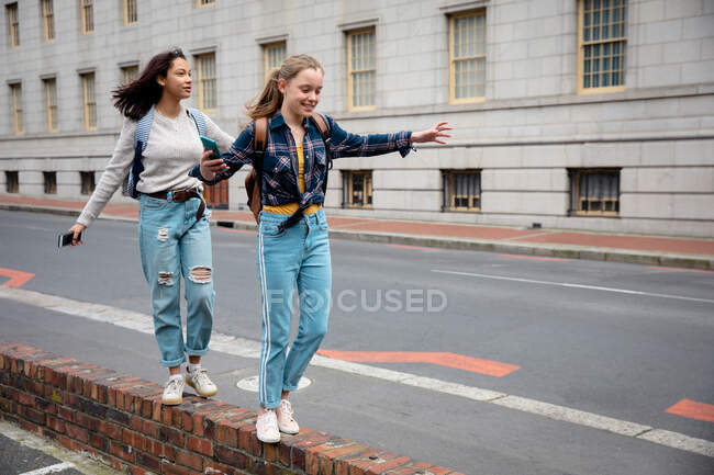 Бічний вид на кавказьку і змішану расу дівчат, які проводять час разом у сонячний день, гуляючи і балансуючи на стіні, посміхаючись. — стокове фото