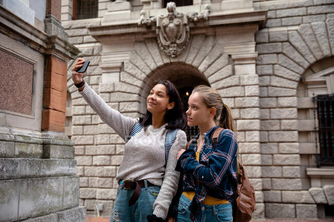 Front view of a Caucasian and a mixed race girls enjoying time hanging out together on a sunny day, standing on the sidewalk, girl taking selfie of herself and her friend. — Stock Photo