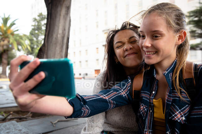 Vue de face d'une Caucasienne et d'une fille métissée profitant du temps passé ensemble par une journée ensoleillée, assise sur un banc, fille prenant selfie d'elle-même et de son amie. — Photo de stock