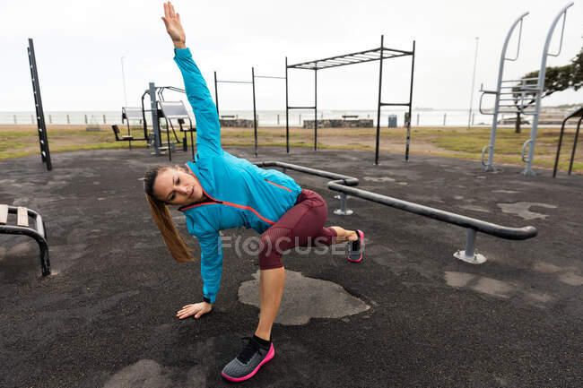 Vista frontal de uma mulher caucasiana desportiva com longos cabelos escuros exercitando-se em um ginásio ao ar livre durante o dia, inclinando-se no chão, alongamento . — Fotografia de Stock