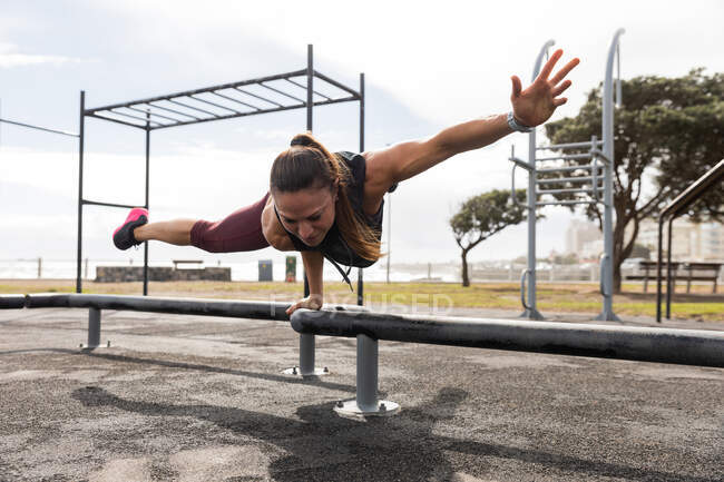 Vista frontal de una mujer atlética caucásica con cabello largo y oscuro haciendo ejercicio en un gimnasio al aire libre durante el día, balanceándose en una mano en un banco. - foto de stock