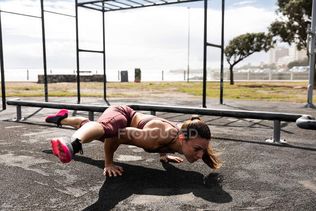 Vista lateral de uma mulher caucasiana desportiva com longos cabelos escuros exercitando-se em um ginásio ao ar livre durante o dia, equilibrando em suas mãos com as pernas estendidas . — Fotografia de Stock
