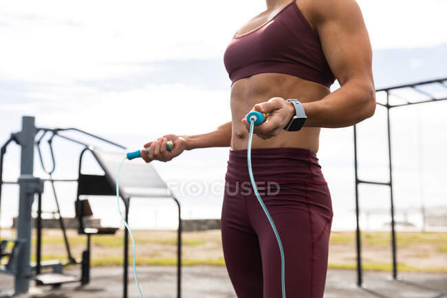 Vista lateral sección media de una mujer deportiva ejercitándose en un gimnasio al aire libre durante el día, sosteniendo una cuerda para saltar. - foto de stock