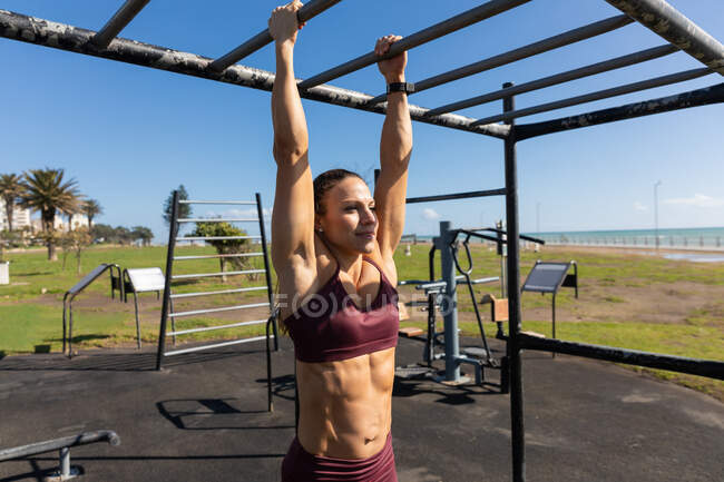 Side view of a sporty Caucasian woman with long dark hair exercising in an outdoor gym during daytime, hanging off an exercising frame. — Stock Photo