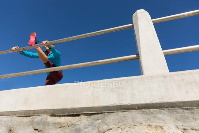 Vista lateral de ángulo bajo de una mujer atlética caucásica con el pelo largo y oscuro ejercitándose en un paseo marítimo en un día soleado con cielo azul, estirando su pierna sobre un pasamanos. - foto de stock