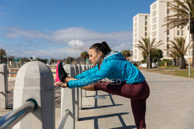 Vista lateral de uma mulher caucasiana desportiva com longos cabelos escuros se exercitando em um passeio à beira-mar em um dia ensolarado com céu azul, esticando a perna em um corrimão . — Fotografia de Stock