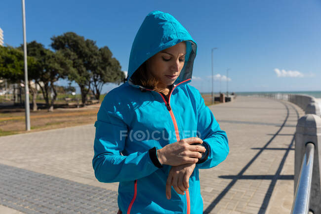 Vista lateral de uma mulher caucasiana esportiva com longos cabelos escuros se exercitando em um passeio à beira-mar em um dia ensolarado com céu azul, verificando seu smartwatch com seu capuz em . — Fotografia de Stock