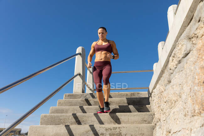 Vista frontal de ángulo bajo de una mujer atlética caucásica con el pelo largo y oscuro haciendo ejercicio en un paseo marítimo en un día soleado con cielo azul, corriendo por las escaleras. - foto de stock
